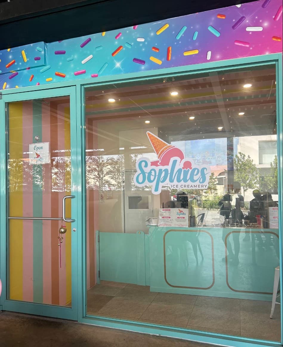 Sophie’s Ice Creamery storefront at 1950 McLeary Ave: a turquoise-framed glass door with pastel stripes and a large window with the Sophie’s logo, under a colorful sprinkle mural