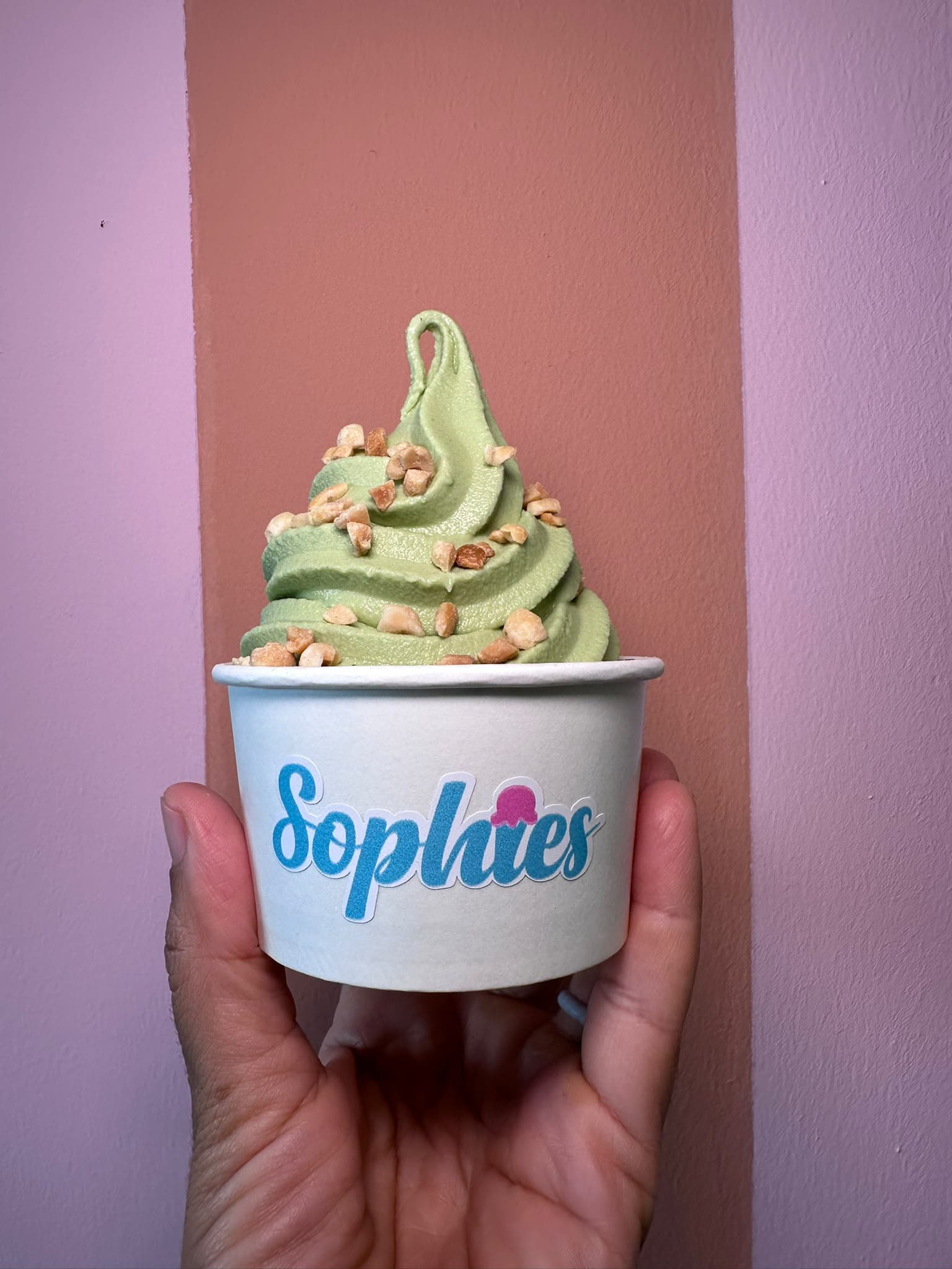 Un swirl de matcha cubierto de maní picado en un vaso de Sophie’s, sostenido frente a una pared terracota y lila