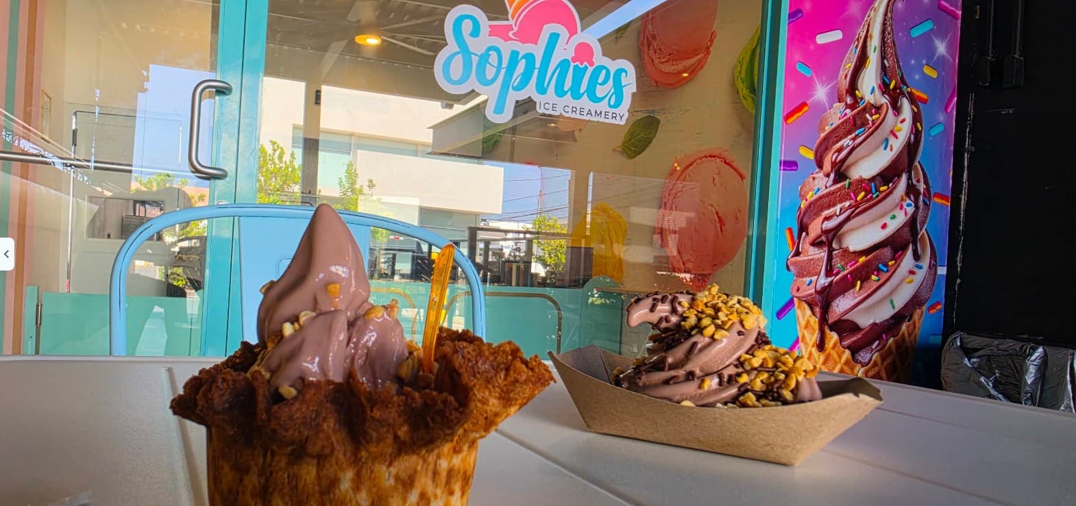 Two treats on a white table at Sophie’s seating area: a chocolate swirl in a waffle bowl and a chocolate swirl with peanuts in a paper tray, in front of the shop window and a mural of a chocolate-and-vanilla cone