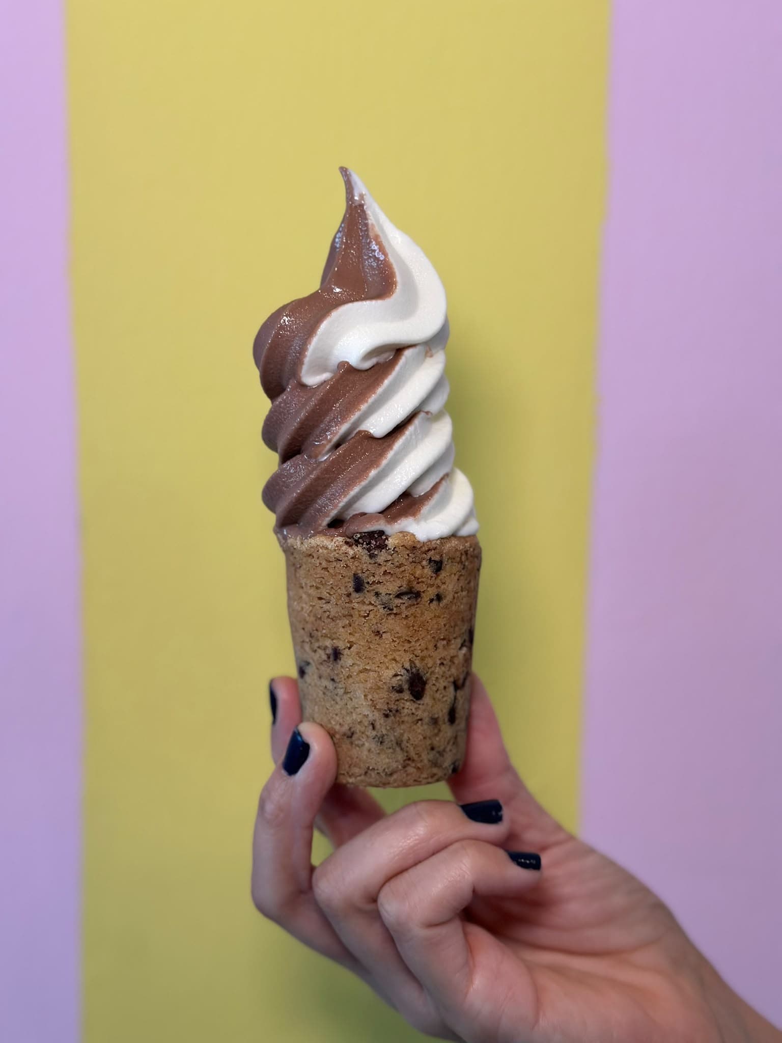 Una mezcla de chocolate y vainilla en un vaso de galleta con chispas de chocolate, sostenido en la mano frente a una pared amarilla y lila