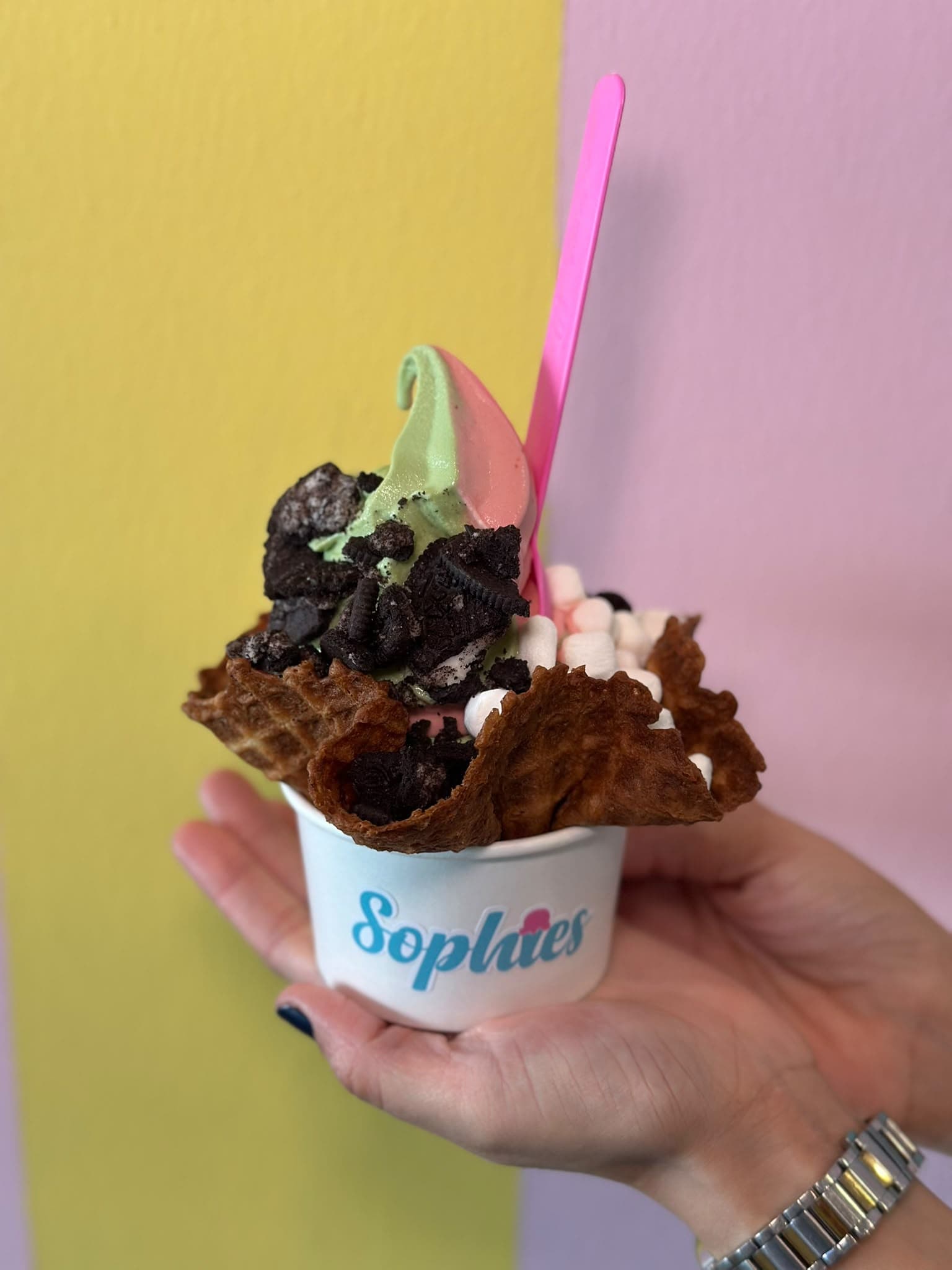 Una mezcla de matcha y fresa en un tazón de waffle crujiente con trocitos de Oreo y mini marshmallows, servida en un vaso de Sophie’s con una cuchara rosada