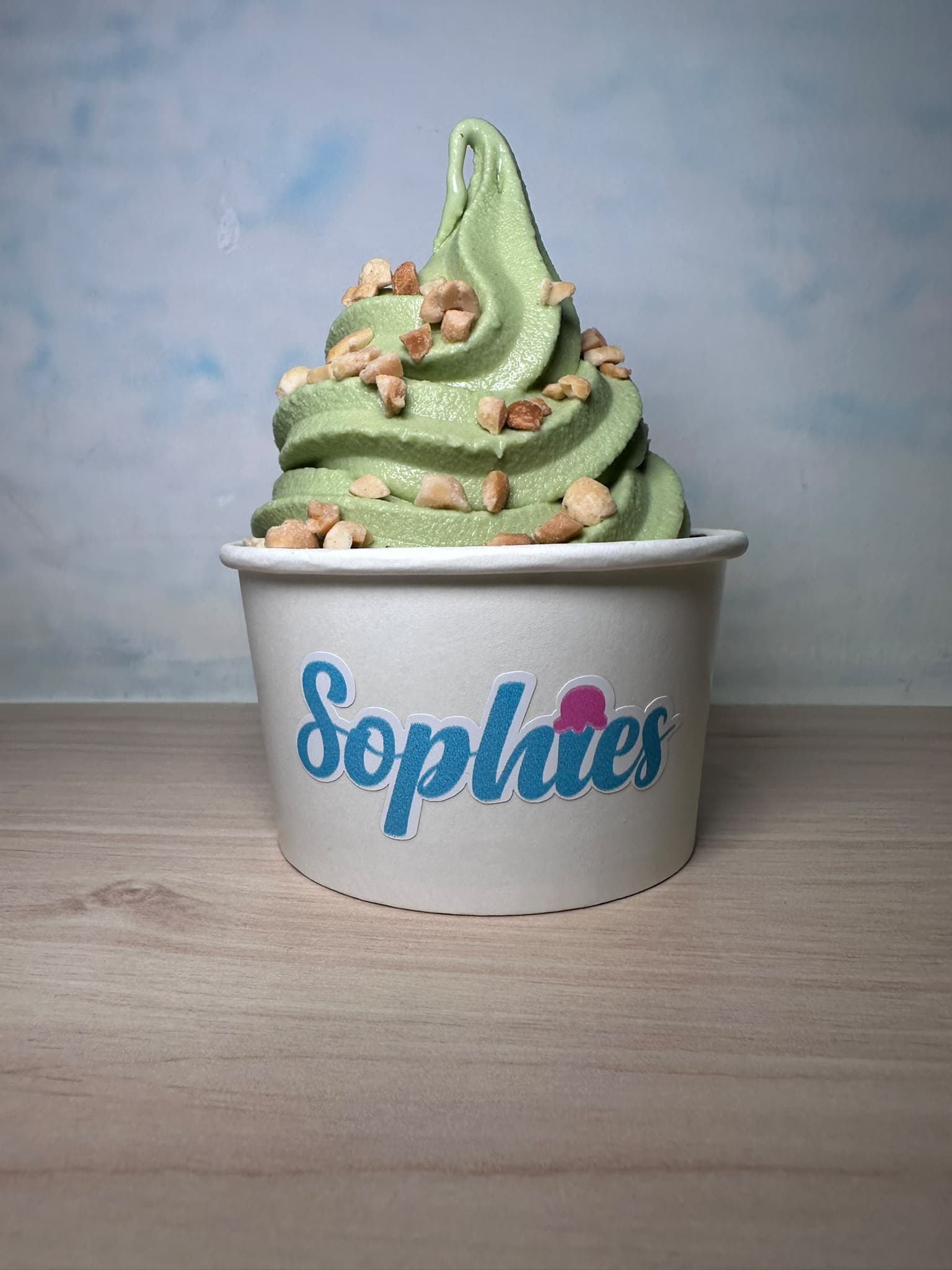 Un swirl de matcha con maní picado en un vaso de Sophie’s sobre una mesa de madera, con un fondo azul cielo suave