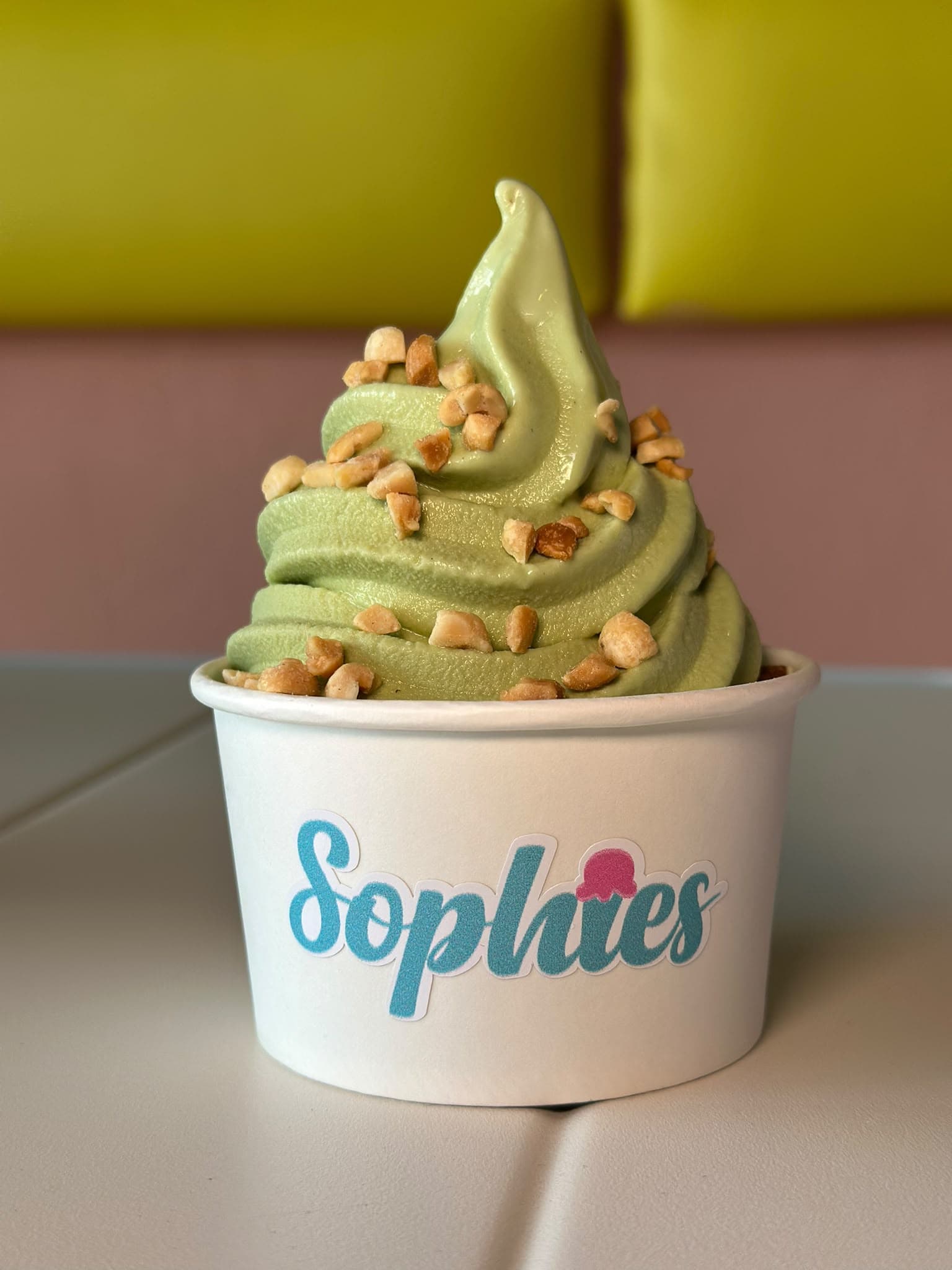 Un swirl de matcha con maní picado en un vaso de Sophie’s sobre una mesa blanca, con una pared amarilla y rosada detrás
